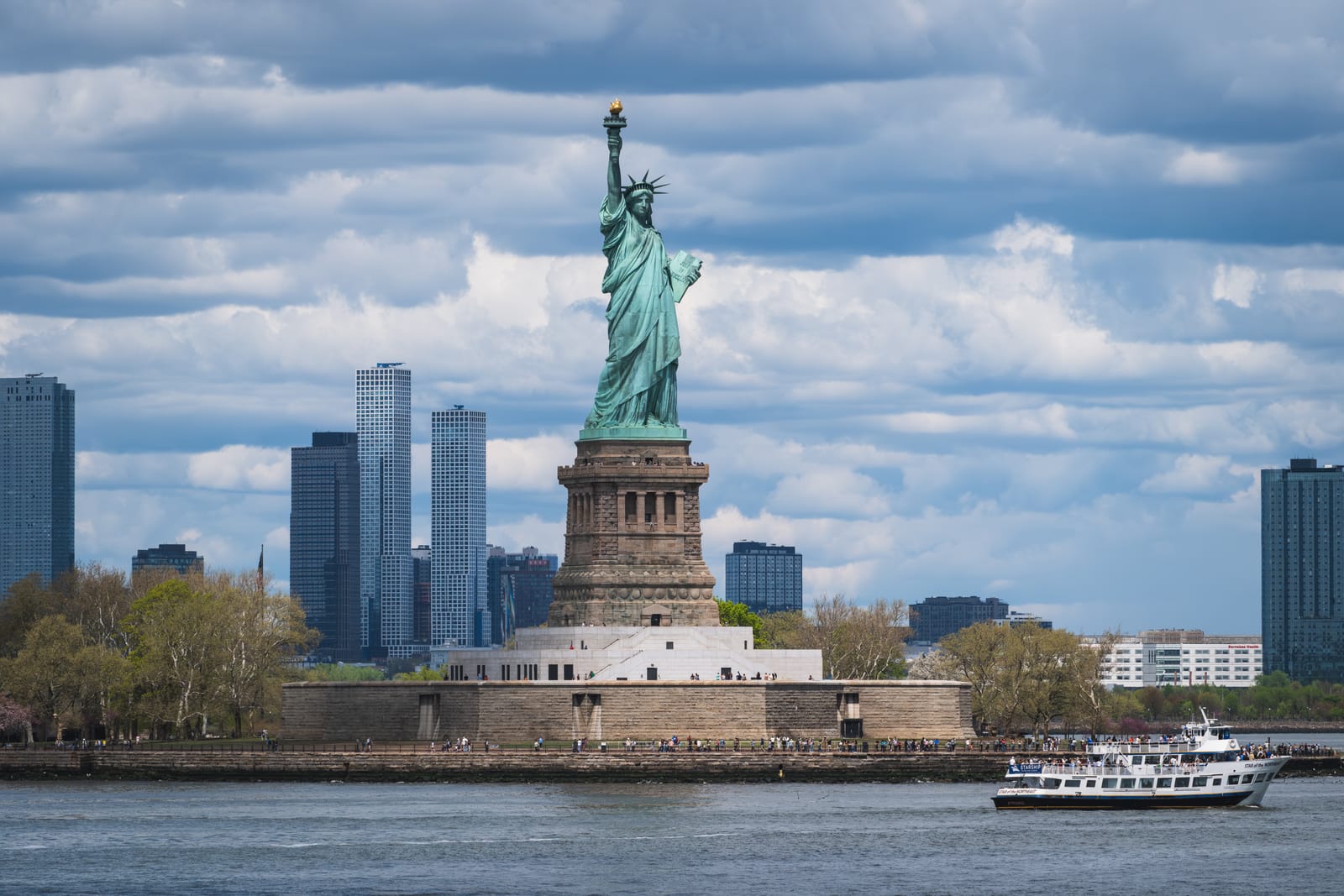 The Statue of Liberty on Liberty Island, seen from the water, with a city skyline behind it